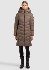 Brązowa puchowa kurtka z pikowanym wzorem, kapturem i zamkiem z przodu. Posiada boczne kieszenie i sięga do połowy uda. Nosi się ją na czarne legginsy i buty.