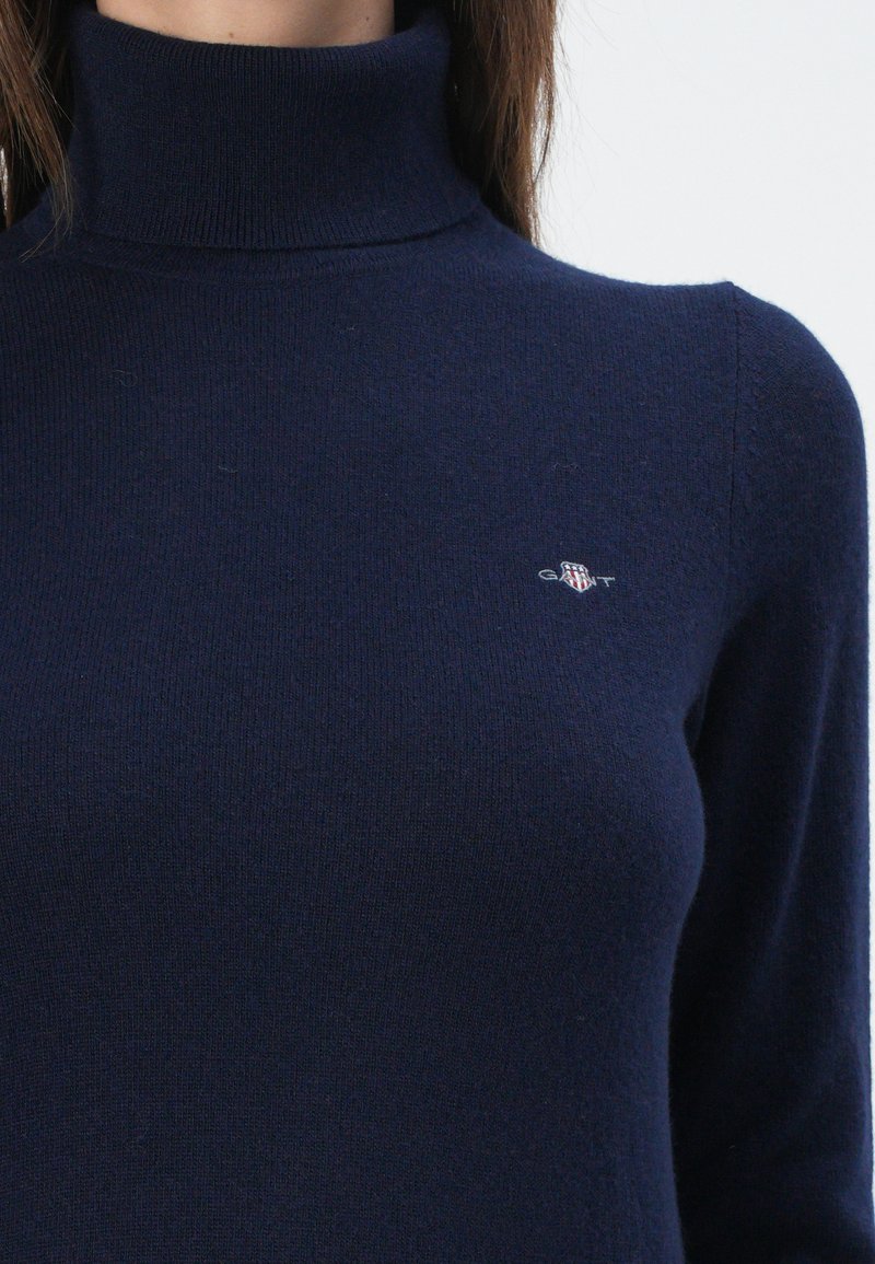 Frau, die einen dunkelblauen Rollkragenpullover trägt, auf dem links auf der Brust ein kleines GANT-Logo gestickt ist.
