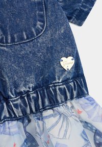 Dżinsowa sukienka z falbaną, przezroczystą, jasnoniebieską warstwą na dole, z serduszkowym, srebrnym logo i detalem w postaci kieszeni.