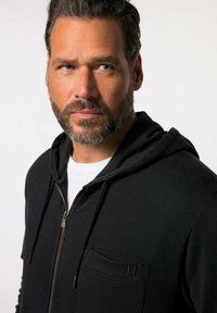 Sudadera con cremallera negra hecha de tela suave, que cuenta con un bolsillo frontal, cordones y un logotipo sutil en el pecho.