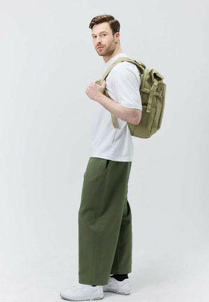 Groene rugzak met verstelbare schouderbanden en meerdere zakken, gedragen over een witte T-shirt en losse groene broek, met witte sneakers.
