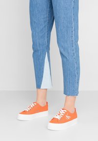 Orange canvas sneakers med vit gummisula, med hål och snörning. Bärs tillsammans med blå denim capribyxor med en ljusblå detalj på sidan.