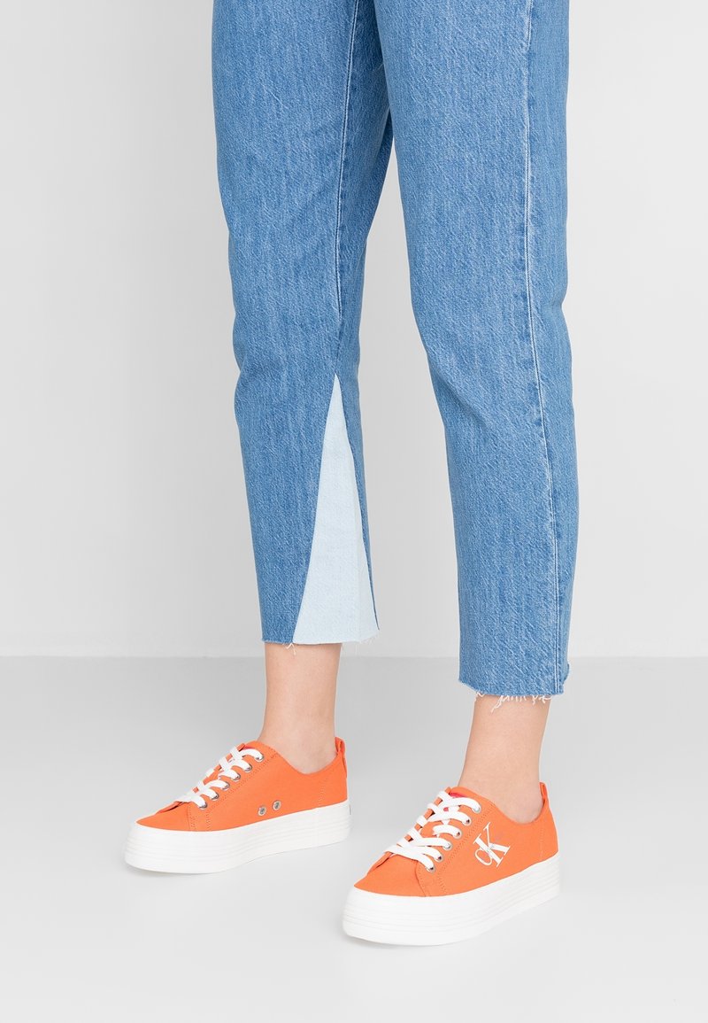 Orange canvas sneakers med vit gummisula, med hål och snörning. Bärs tillsammans med blå denim capribyxor med en ljusblå detalj på sidan.