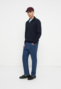 Marine blauw rits sweatshirt, lichtblauw shirt eronder, donkere spijkerbroek, zwarte schoenen en een bordeauxrode baseballpet naar voren gedragen. Eenvoudig ontwerp, relaxte pasvorm.