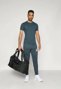 Sportoutfit mit einem eng anliegenden, dunkelgrünen Kurzarmshirt und passenden Jogginghosen mit einem weißen Logo, kombiniert mit einer großen schwarzen Sporttasche.