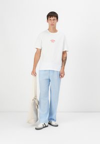 White cotton t-shirt with red graphic text, light blue linen pants, gray shoes with black stripes, and a beige tote bag. Simple design.