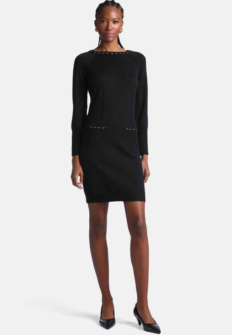 Femme debout portant une robe en maille noire à manches longues avec des poches et des talons noirs sur un fond blanc uni.