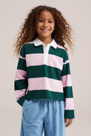 Jeune fille souriante aux cheveux bouclés portant un t-shirt à manches longues rayé vert et rose et un pantalon bleu clair sur un fond uni.