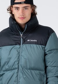 Veste rembourrée en vert foncé et noir avec un col haut, fermeture zippée et logo Columbia sur la poitrine. Fabriquée en tissu brillant et matelassé.