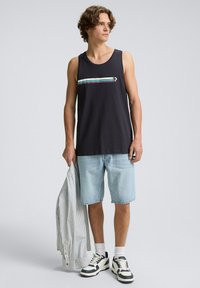 Top senza maniche nero con strisce orizzontali multicolori. Pantaloni corti in denim chiaro, calze bianche e scarpe da ginnastica in bianco e nero. Camicia a righe in mano.