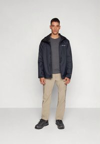 Men's black waterproof jacket with a hood, gray shirt, beige cargo pants, and black hiking boots. Smooth fabric and practical design.