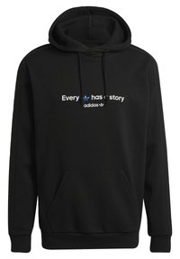 Sort hættejakke lavet af blødt materiale med en kængurulomme. Foran står teksten "Alle har en historie" med et blåt Adidas-logo ovenfor og nedenfor.