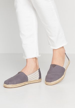 Stopy w szarych espadrylach typu slip-on z plecionymi jucianymi podeszwami i białymi paskowanymi bokami z tkaniny, zestawione z białymi spodniami z postrzępionym dołem.
