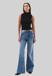 Top negro sin mangas, con cuello alto; jeans de pierna ancha de color azul claro con un cinturón con detalles en plata; modelo de pie contra un fondo gris.