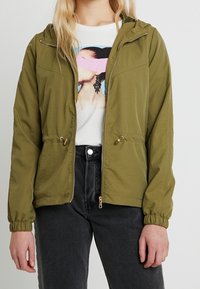 Olivenfarvet letvægtsjakke med hætte, lynlås foran, justerbare snore i taljen og elastiske håndled. Båret over en grafisk T-shirt.