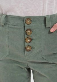 Pantalon en tissu vert à taille haute, avec quatre boutons marron sur le devant. Le matériau semble doux avec une texture lisse.