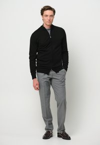 Maglione zip-up nero con un design aderente, indossato sopra una camicia grigia. Abbinato a pantaloni leggeri grigi e mocassini marrone scuro.