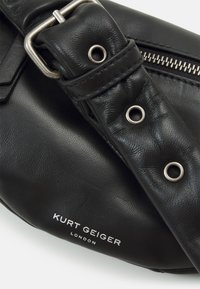 Musta nahkainen käsilaukku, jossa on sileä pinta, hopeanväriset metalliosat, rei'ittäjät ja etuosan vetoketju. Laukussa näkyy "KURT GEIGER LONDON" -logo.