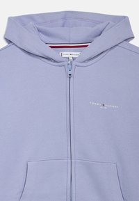 Lyse lilla zip-up hettegenser laget av mykt stoff, med en frontlomme, hette, og en liten merke-logo på venstre bryst.