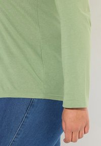 Světle zelené tričko s dlouhým rukávem, kulatým výstřihem a rovým lemem, v kombinaci s modrými džíny. Hladká a jemná textilie.