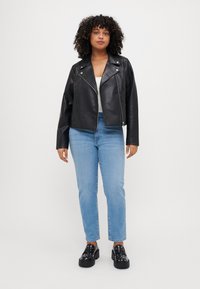 Göndör hajú nő fekete bőrdzsekiben, fehér felsőben, világos kék farmernadrágban és fekete tüskés platform cipőkben áll egy fehér háttér előtt.