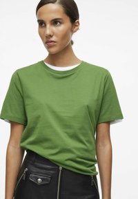 Camiseta de algodón verde con cuello redondo y mangas cortas, superpuesta a una camisa blanca. Llevada con una falda negra texturizada con cremalleras.