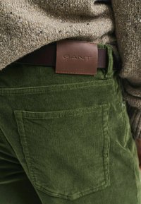 Acercamiento de pantalones de pana verdes con un bolsillo trasero y una etiqueta de cinturón de cuero marrón con la inscripción "GANT" debajo de un suéter de punto beige.