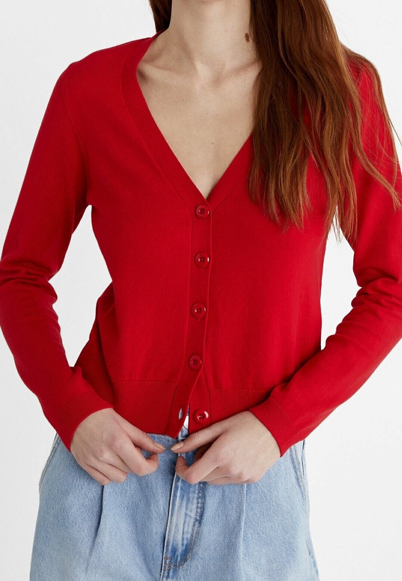 Cardigan rouge avec un col en V, manches longues et six boutons sur le devant. Confectionné en tissu tricoté doux. Associé à un jean en denim bleu clair.