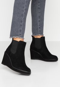 Bottines à talons compensés en suédine noire avec des panneaux élastiques sur les côtés, bout rond et coutures visibles le long de la semelle. Portées avec un jean gris à ourlet effiloché.