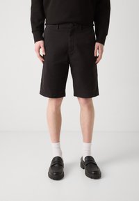Shorts en coton noir avec une coupe droite, passants de ceinture et fermeture à bouton. Portés avec des chaussettes blanches et des chaussures noires à enfiler.