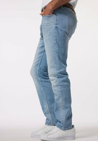 Vaqueros de denim azul claro con un diseño ajustado, detalles sutiles de desgaste, cinco bolsillos y una cinturilla lisa, combinados con zapatillas blancas.