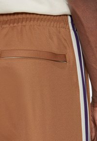 Primer plano de una persona que lleva pantalones de chándal marrones con cinturilla elástica, bolsillo trasero con abertura y franjas laterales blancas y moradas.