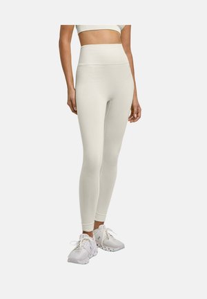 Vrouw die crèmekleurige leggings met hoge taille en witte sportschoenen draagt, staand tegen een effen lichte achtergrond.