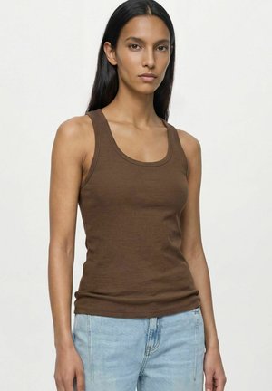 Femme aux longs cheveux foncés porte un débardeur marron sans manches et un jean bleu clair, debout devant un fond clair uni.
