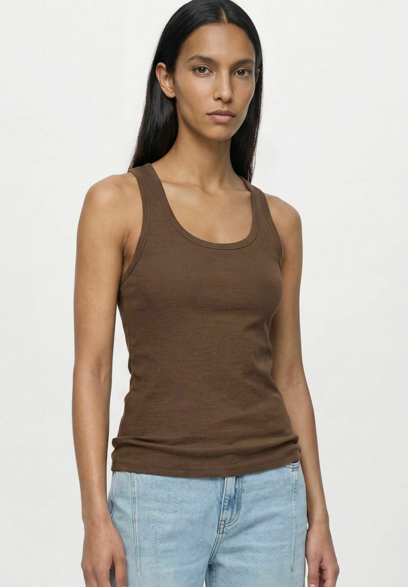 Femme aux longs cheveux foncés porte un débardeur marron sans manches et un jean bleu clair, debout devant un fond clair uni.