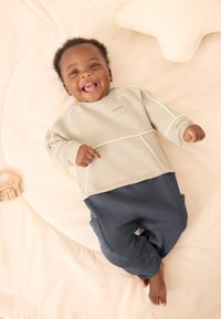 Lysegrøn langærmet top med hvide detaljer og et logo, kombineret med mørkeblå joggingbukser. Barn ligger på en blød, rund, lysefarvet overflade.