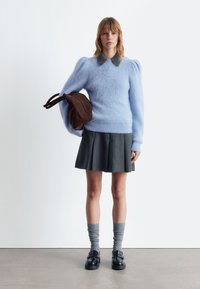 Himmelblauer, flauschiger Pullover mit Puffärmel, darunter ein graues Hemd mit Kragen, kombiniert mit einem grauen Faltenrock und schwarzen Schuhen. Eine braune Handtasche wird gehalten.