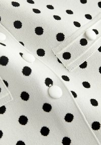 Strikket top i hvid med sorte polkaprikker, med knapdetaljer og en tekstureret stof. Karakteristiske, jævnt fordelte mønstre over hele.