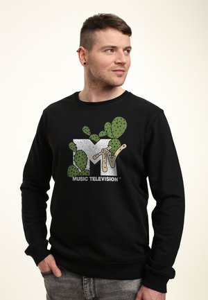 Mand med kort hår og ørepiercinger iført en sort sweatshirt med et grønt kaktusmotiv på MTV-logoen, der kigger til siden.