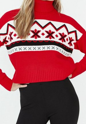 Femme portant un pull à col roulé rouge avec des motifs géométriques blancs et noirs et un pantalon noir taille haute, mains sur les hanches.
