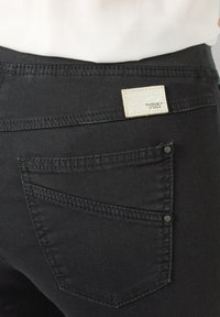 Achteraanzicht van een zwarte spijkerbroekzak met dubbele stiksels en een rechthoekig lichtbeige merklapje met de tekst "Raphaela by Brax."