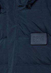 Marinblå pufferjacka med en texturerad yta, med en rektangulär svart gummilogo patch märkt "POLO CLUB."