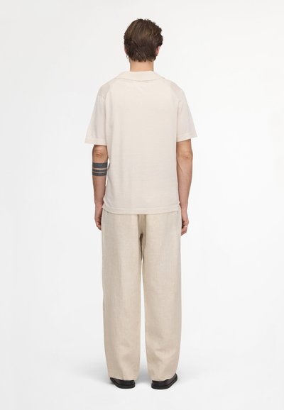 Uomo in piedi di spalle, indossa una camicia beige a maniche corte, pantaloni beige ampi, scarpe nere e un tatuaggio braccialetto nero sull'avambraccio sinistro.