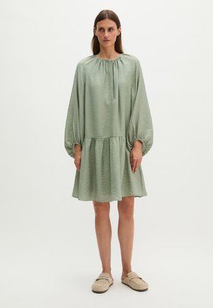 Femme portant une robe ample vert sauge avec des manches ballon et des chaussures beige sans lacets, debout devant un fond clair uni.
