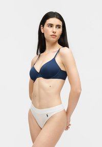 Γυναίκα με μακριά σκούρα μαλλιά, που φοράει μπλε σουτιέν και λευκά εσώρουχα, στέκεται με τα χέρια πίσω από την πλάτη, κοιτώντας προς τα πλάγια σε απλό φόντο.