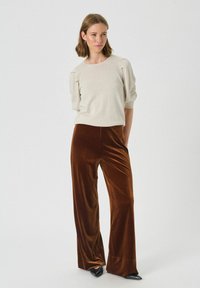 Pulover bej, texturat cu manșete puf, asortat cu pantaloni din catifea maro, cu talie înaltă și largi. Pantofi negri cu vârf ascuțit finalizează ținuta.