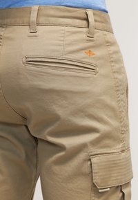 Pantalones cargo beige con una textura suave, que cuentan con dos bolsillos laterales y un bolsillo trasero. Detalle del logo naranja pequeño cerca de la cinturilla.