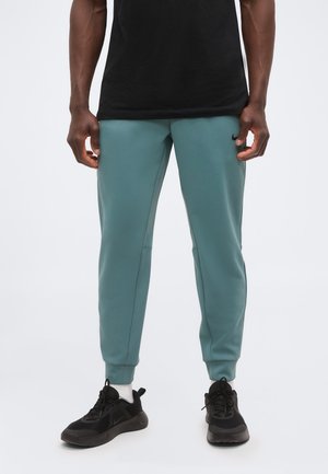 Homme portant un pantalon de jogging vert bleu clair, des baskets noires et des chaussettes blanches, debout devant un fond uni clair.