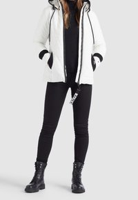 Veste matelassée blanche avec capuche, accents noirs sur les manches et fermeture éclair à l'avant. Le modèle porte des jeans skinny noirs et des bottes de combat noires.