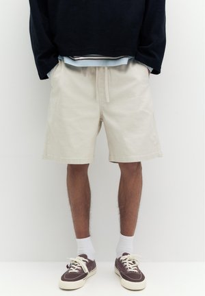 Uomo che indossa pantaloncini beige con coulisse, calzini bianchi corti e sneakers in tela marrone scuro, in piedi davanti a uno sfondo bianco semplice.